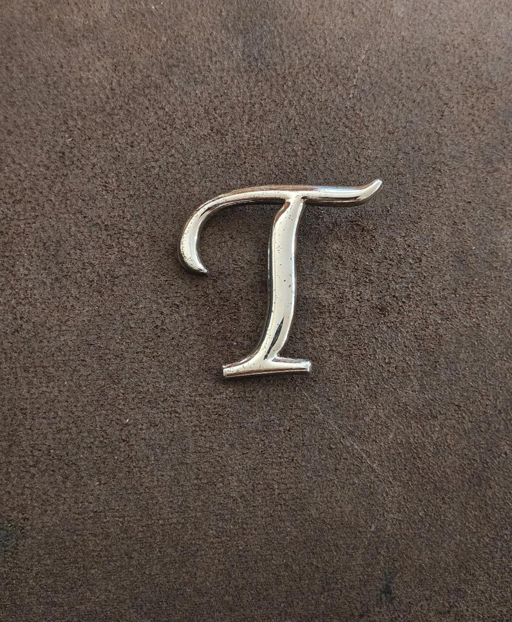 Silver-tone letter T pin/brooch, front view on dark textured background.