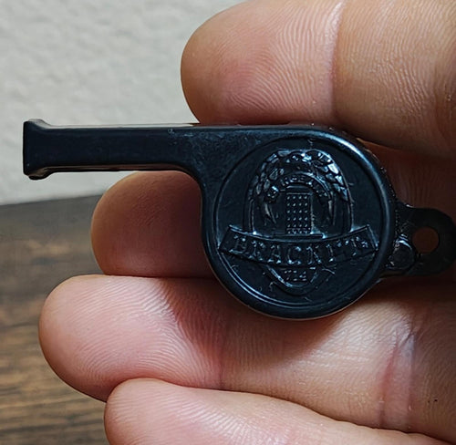 Black whistle with round engraved face and hanging loop, held between fingers.