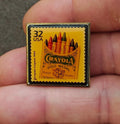Square gold-tone pin with yellow stamp design, “32 USA” and “Crayola Gold Medal”