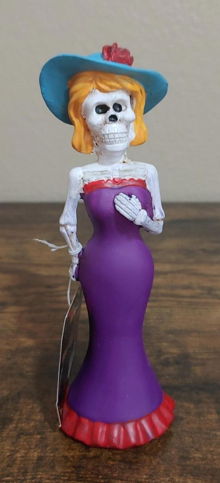 Purple Catrina figurine with blue hat and white skull face, holding a plate
