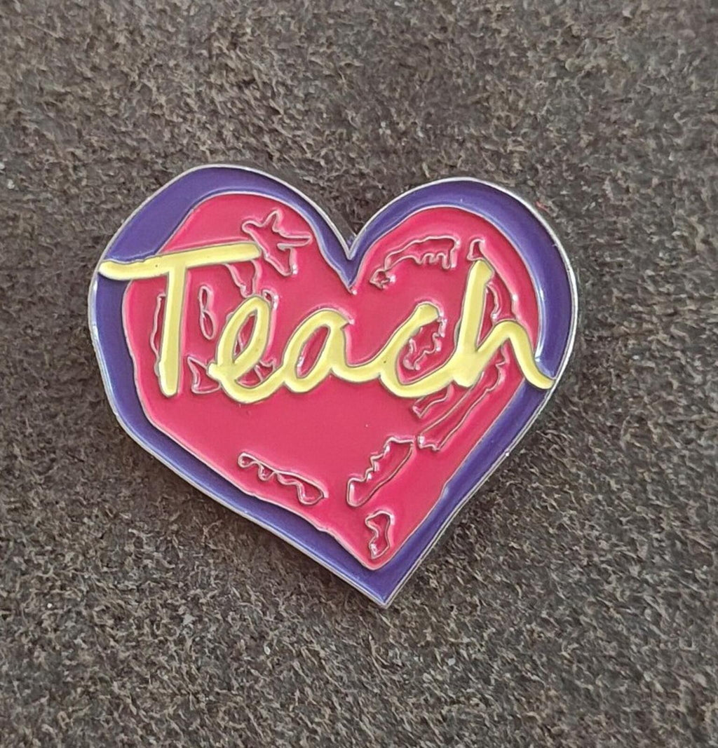 Enamel heart pin on fabric, red “Teach” lettering with yellow strokes inside purple border.