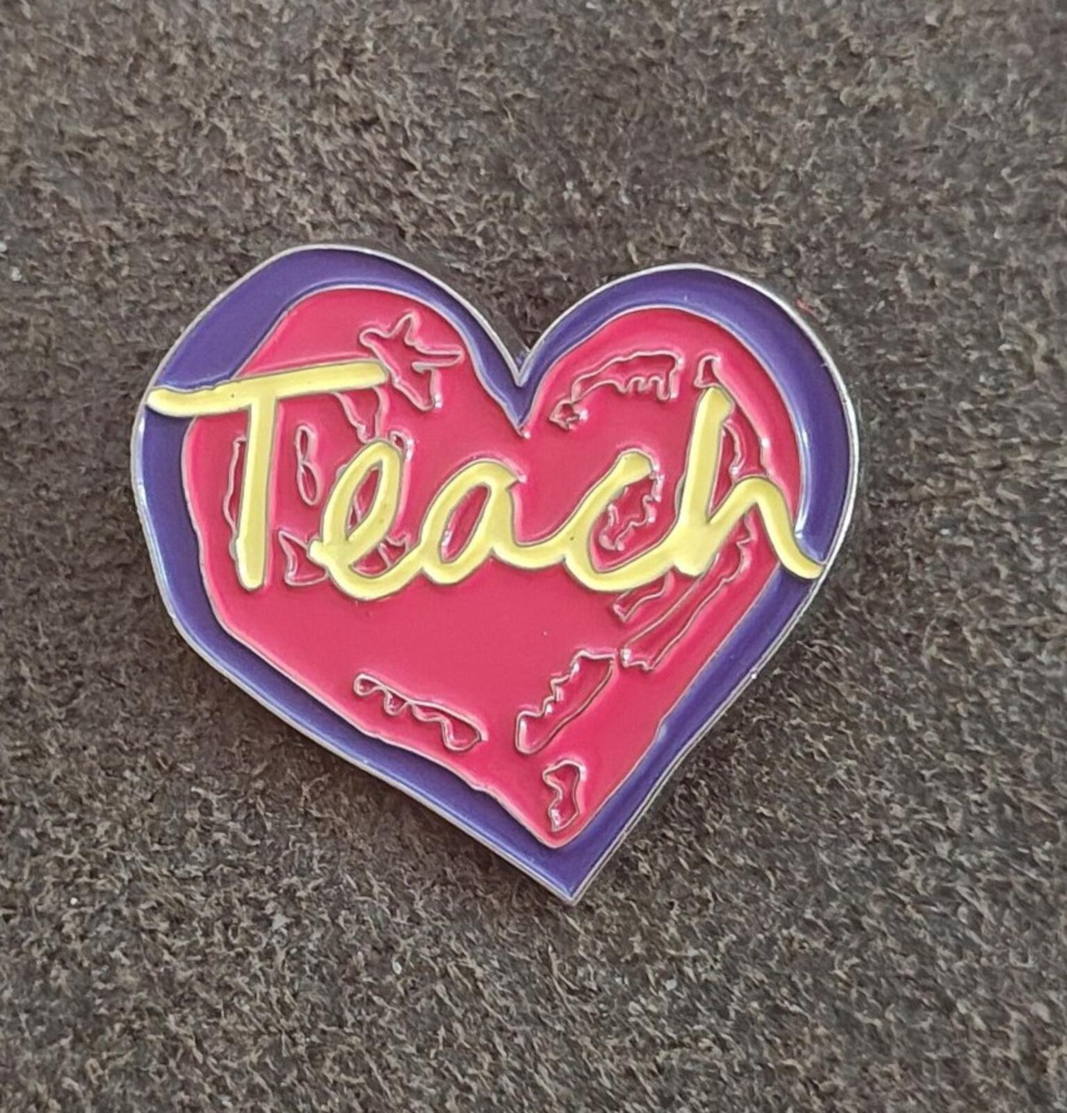 Enamel heart pin on fabric, red “Teach” lettering with yellow strokes inside purple border.