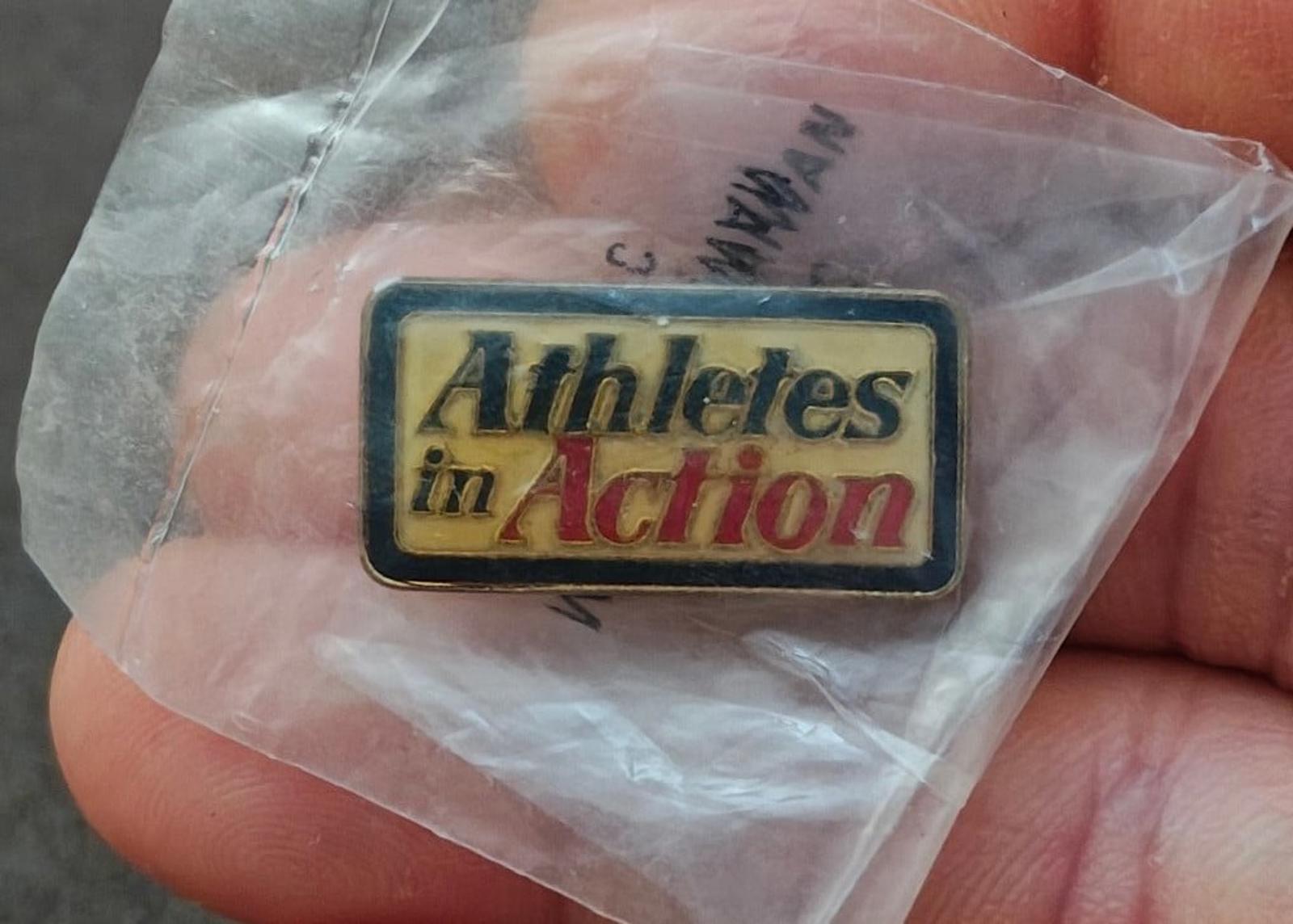 Gold-tone enamel pin "Athletes in Action" close-up with red/yellow lettering, in bag