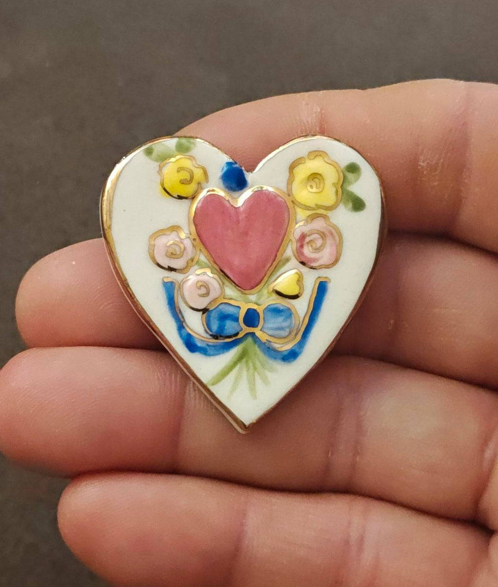 Heart-shaped ceramic brooch pin held in fingers, pink heart floral design with gold-tone edge