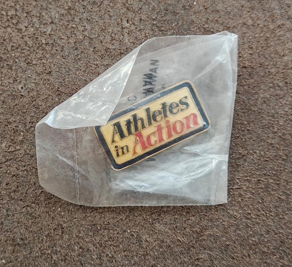 Gold-tone enamel pin reading "Athletes in Action" inside clear bag on gray surface