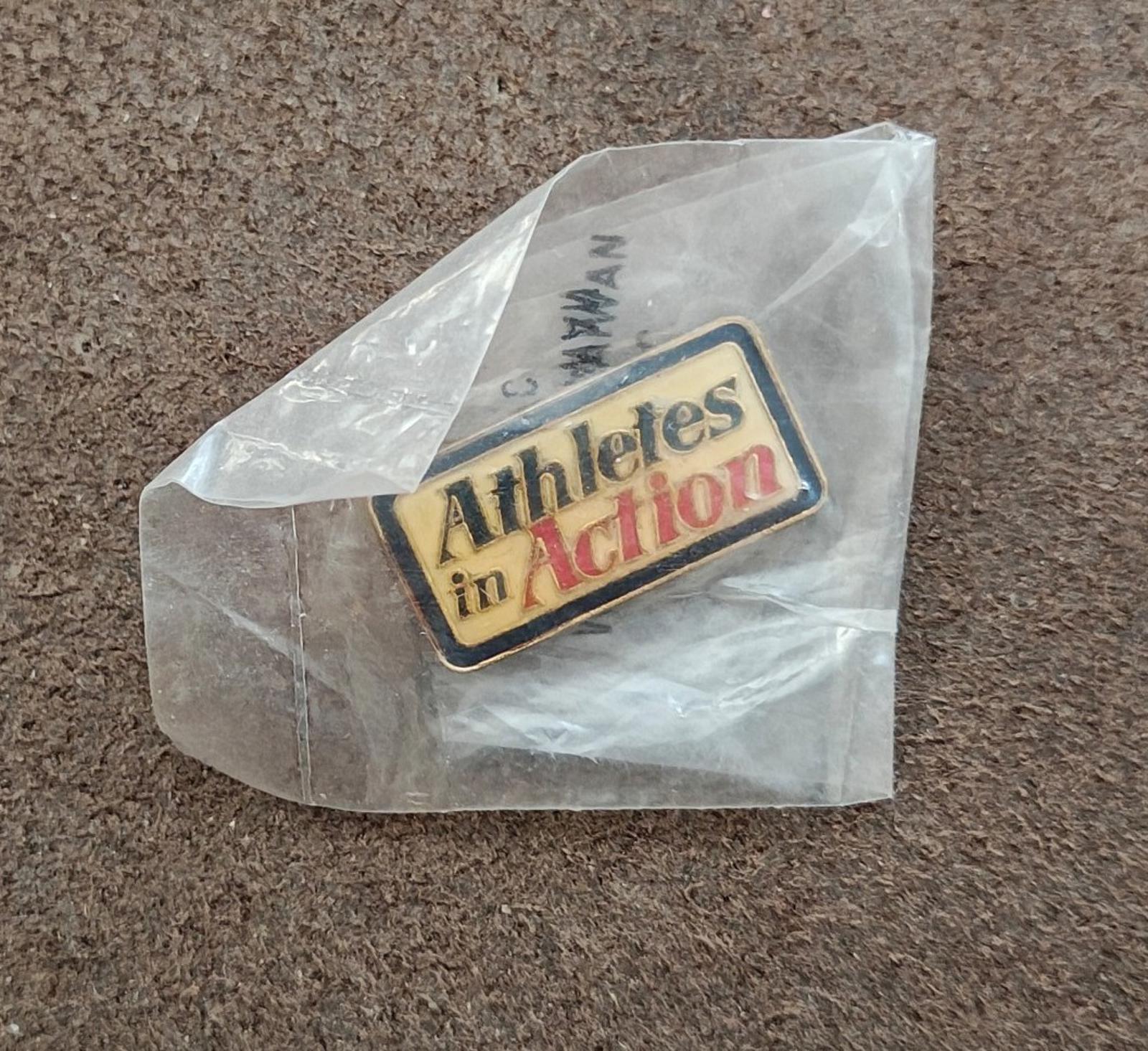 Gold-tone enamel pin reading "Athletes in Action" inside clear bag on gray surface