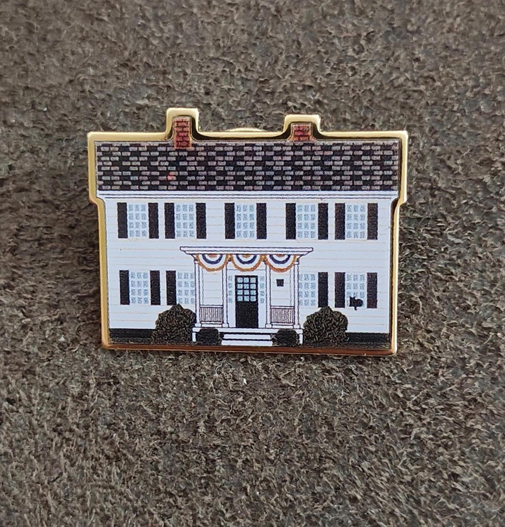 Gold-edged enamel pin with white building facade and black trees, flat on gray fabric