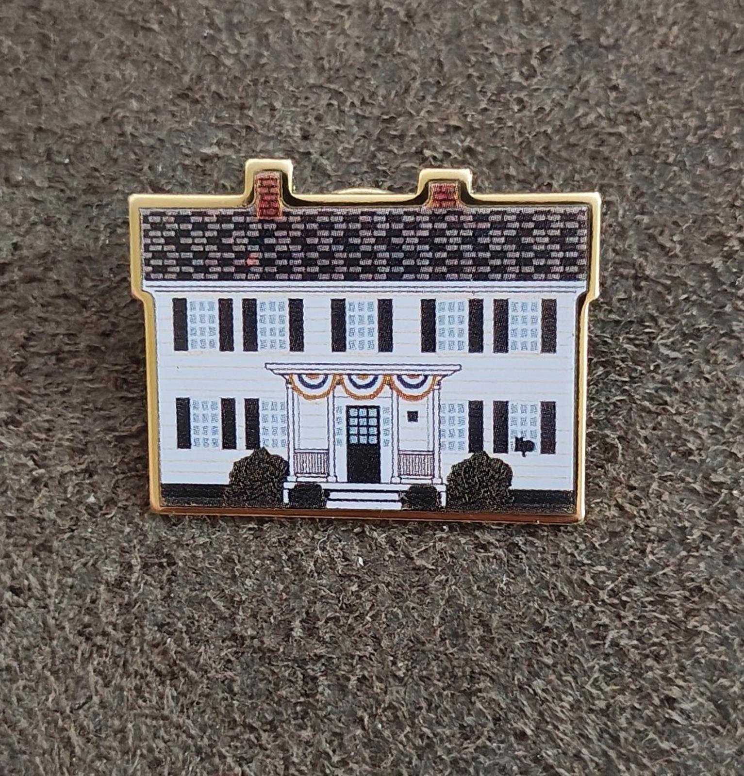 Gold-edged enamel pin with white building facade and black trees, flat on gray fabric