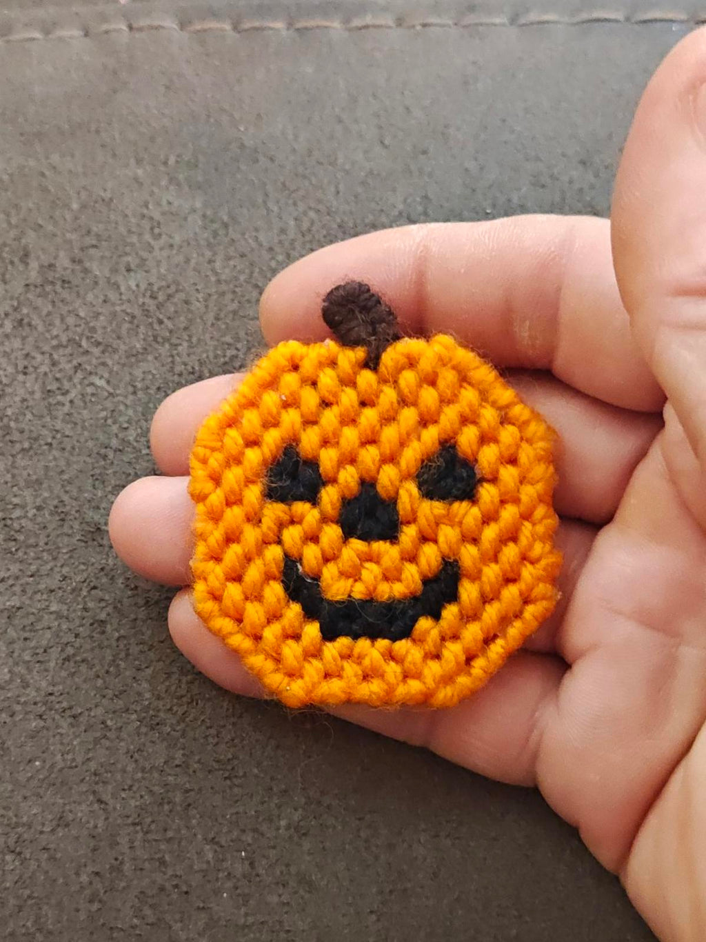 Orange jack o' lantern brooch held in hand, crocheted texture with black smile detail.
