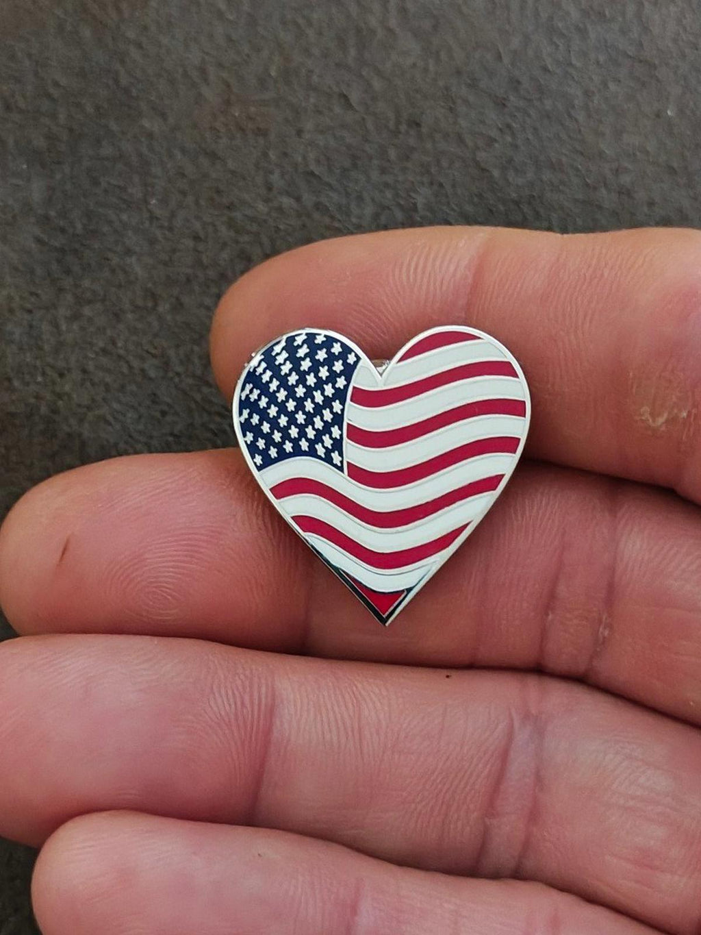 Heart-shaped American flag pin held in fingers, blue stars with red and white wavy stripes