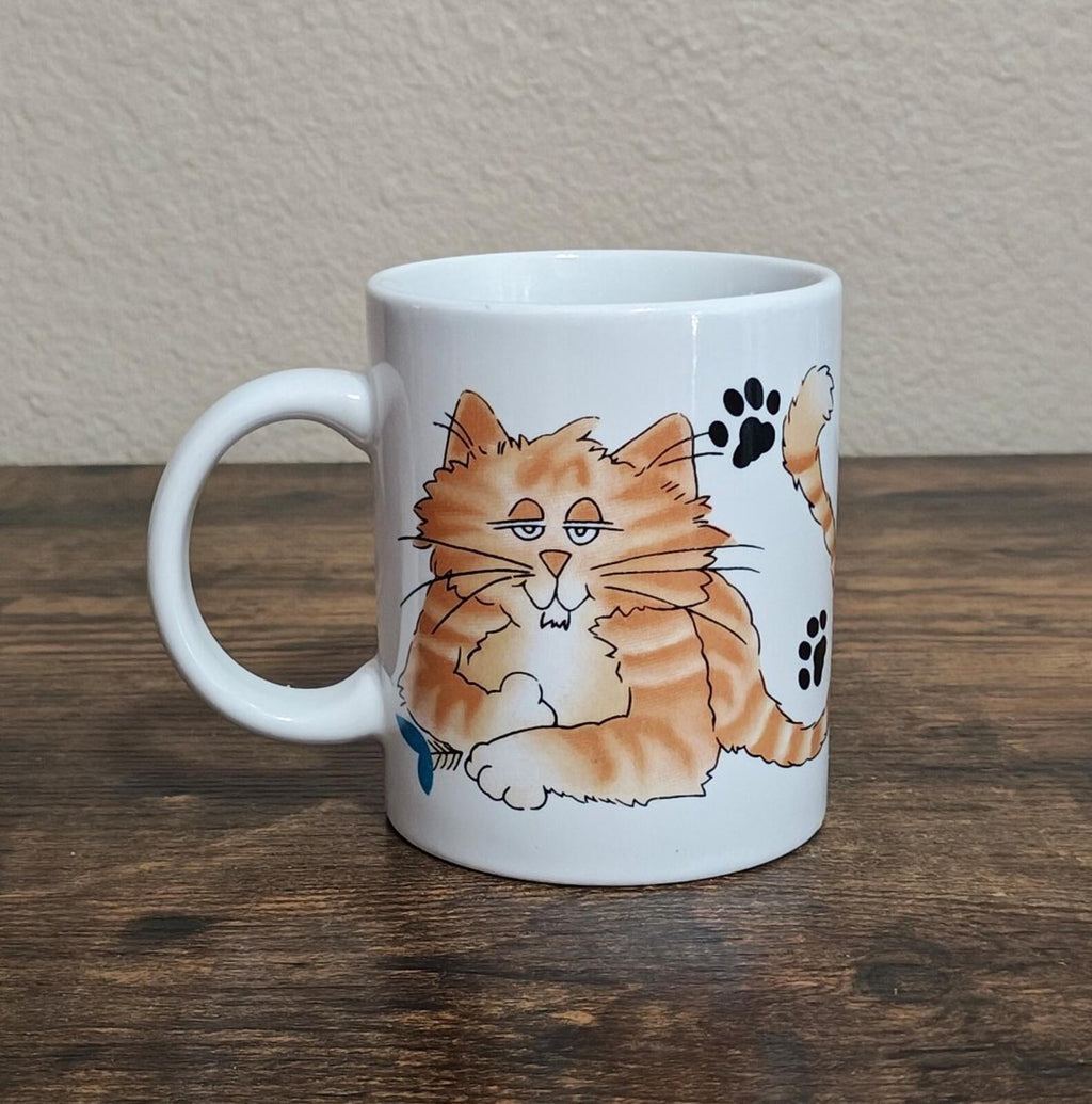 White ceramic mug with orange calico cat face and paw prints, front view on table