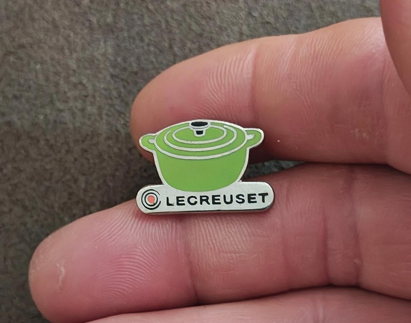 Le Creuset green mini cocotte enamel pin held in fingers, front detail