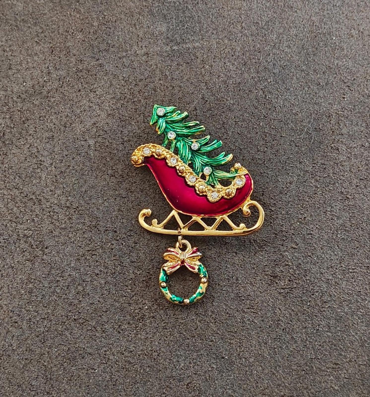 Full view of red and green Christmas sleigh enamel pin with gold runners and wreath dangle.