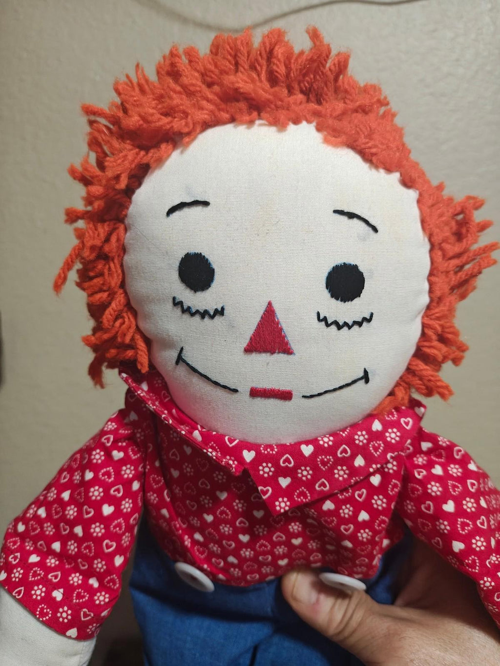 Close-up handmade rag doll face with orange yarn hair, red heart-pattern shirt collar.