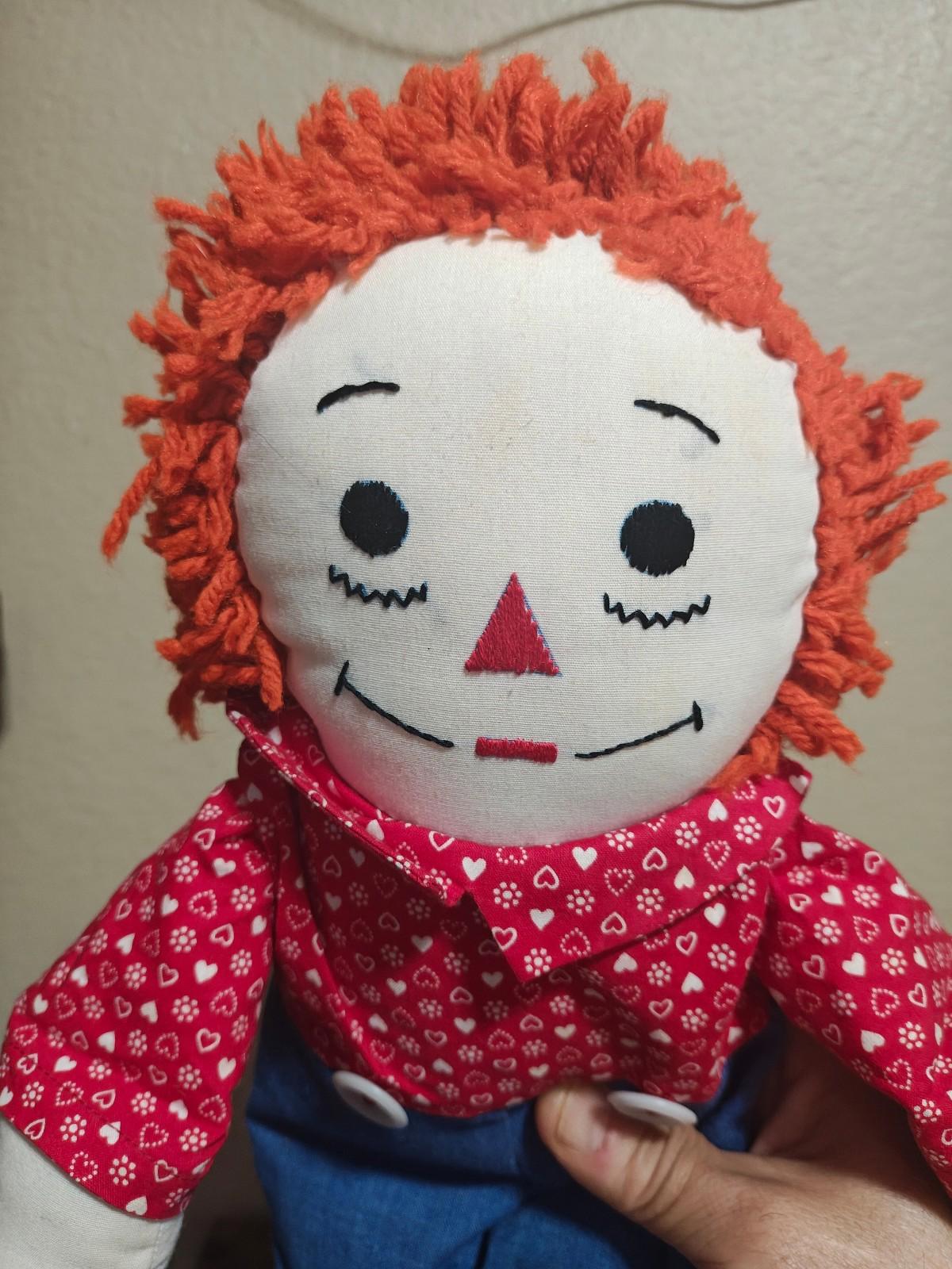 Close-up handmade rag doll face with orange yarn hair, red heart-pattern shirt collar.