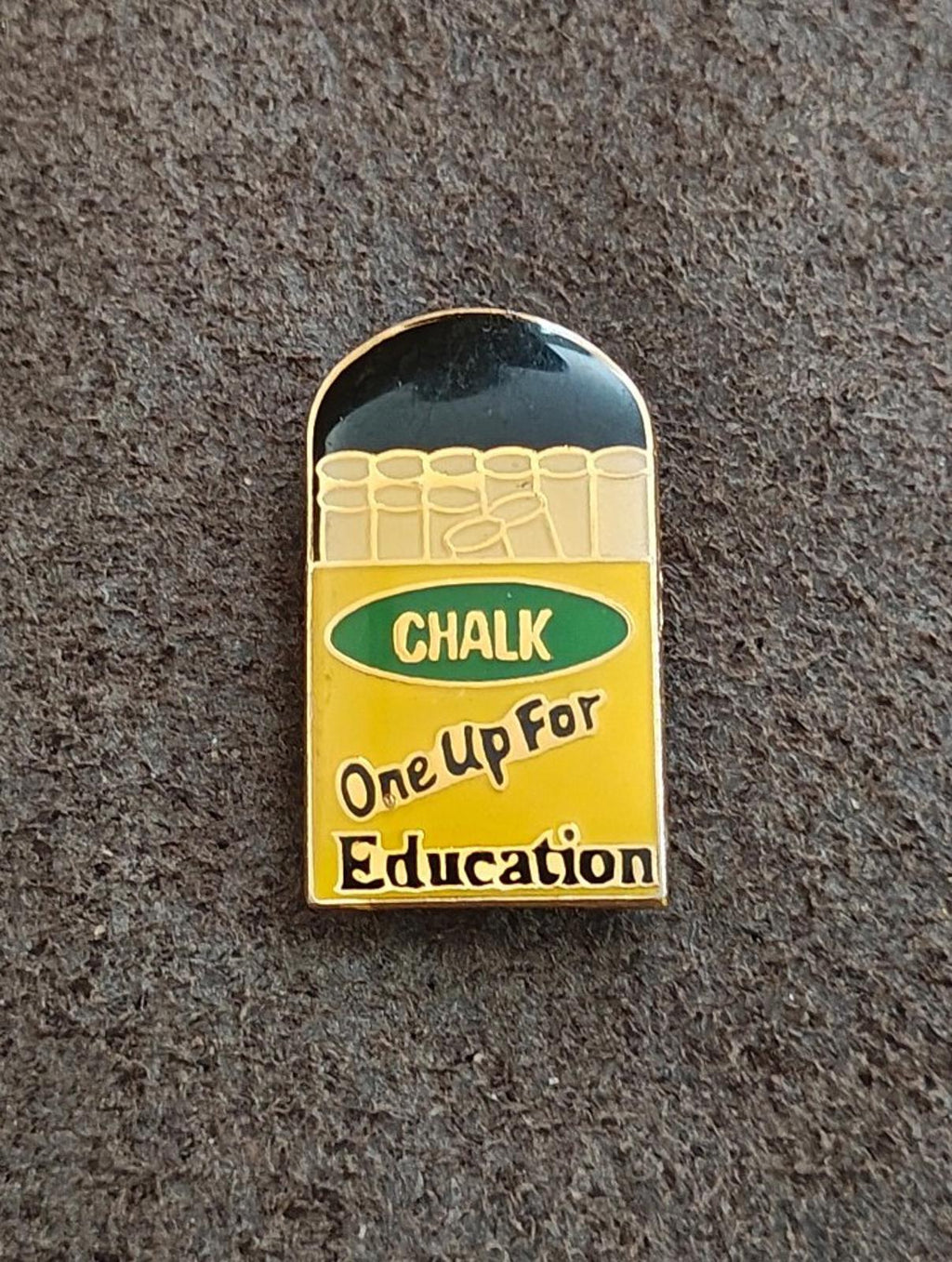 Gold-tone enamel teacher pin on gray surface, chalkboard “CHALK” and “One Up For Education”