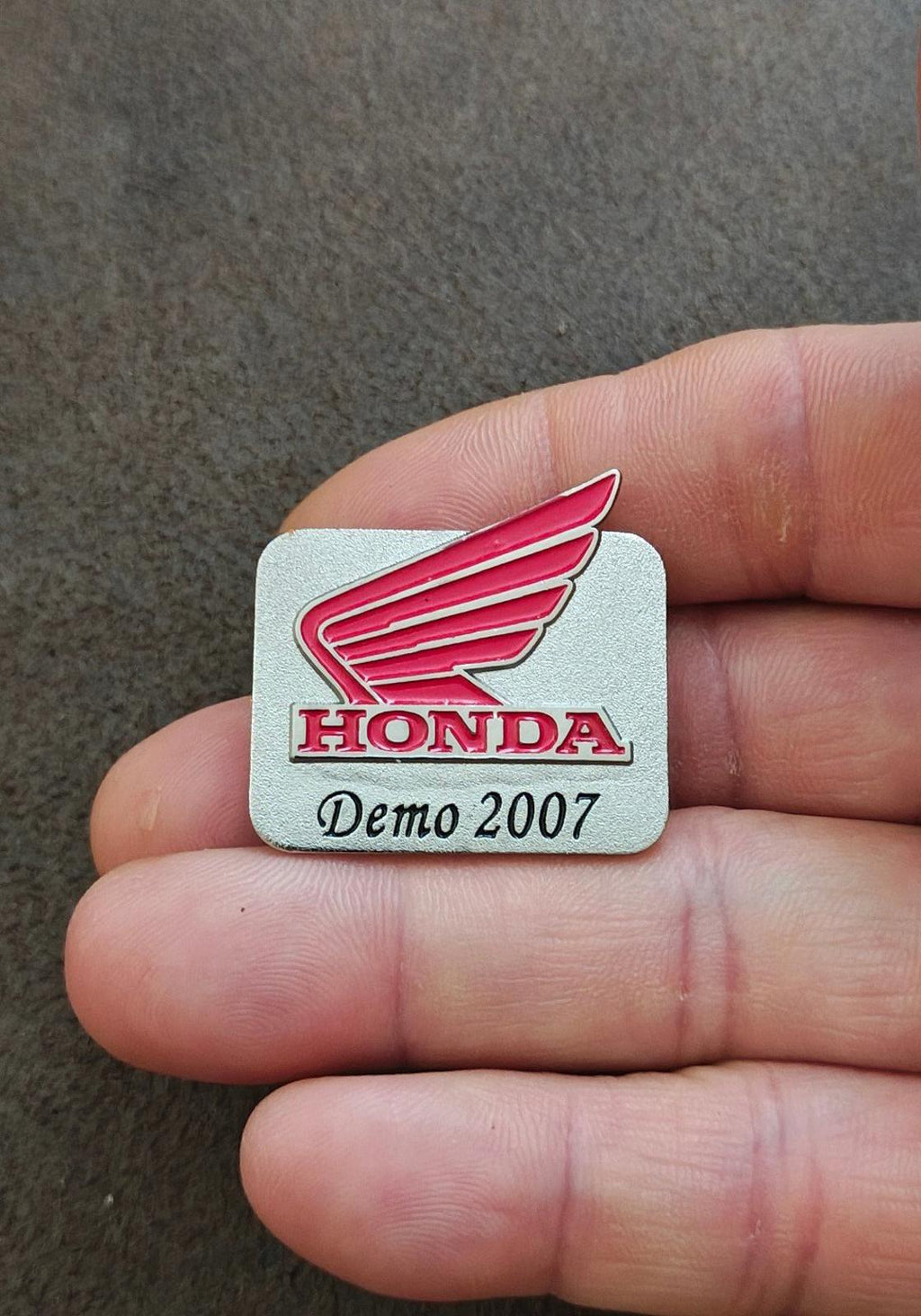 Silver-tone Honda Demo 2007 enamel pin in hand view, red wing logo and HONDA