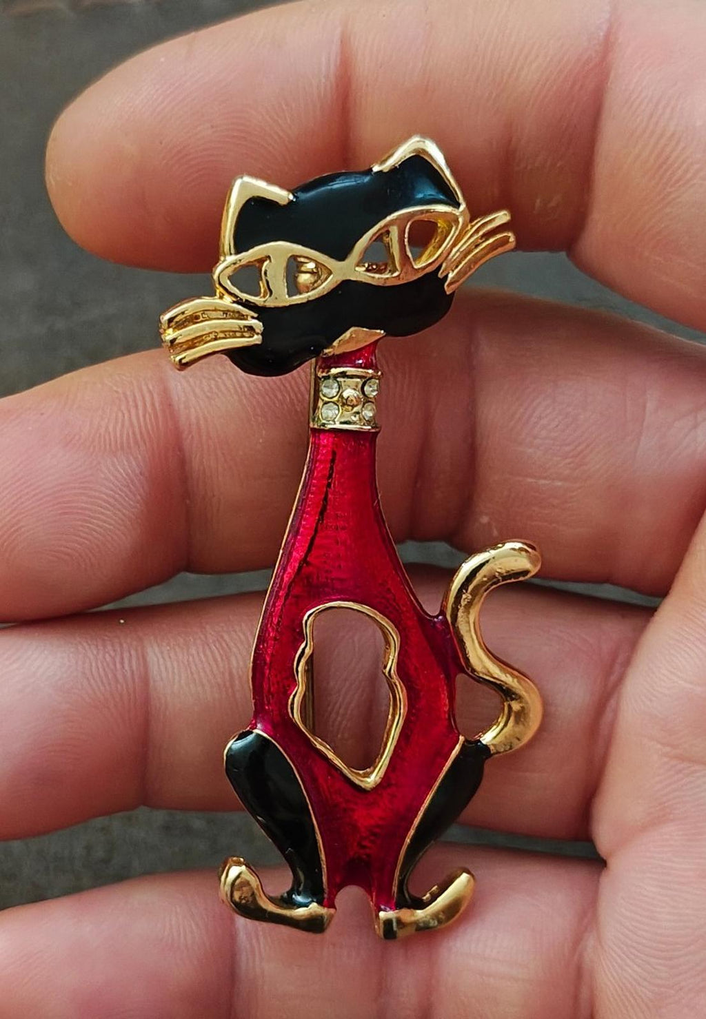 Red and black enamel cat mask brooch with gold-tone detailing, held in fingertips.