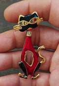 Red and black enamel cat mask brooch with gold-tone detailing, held in fingertips.
