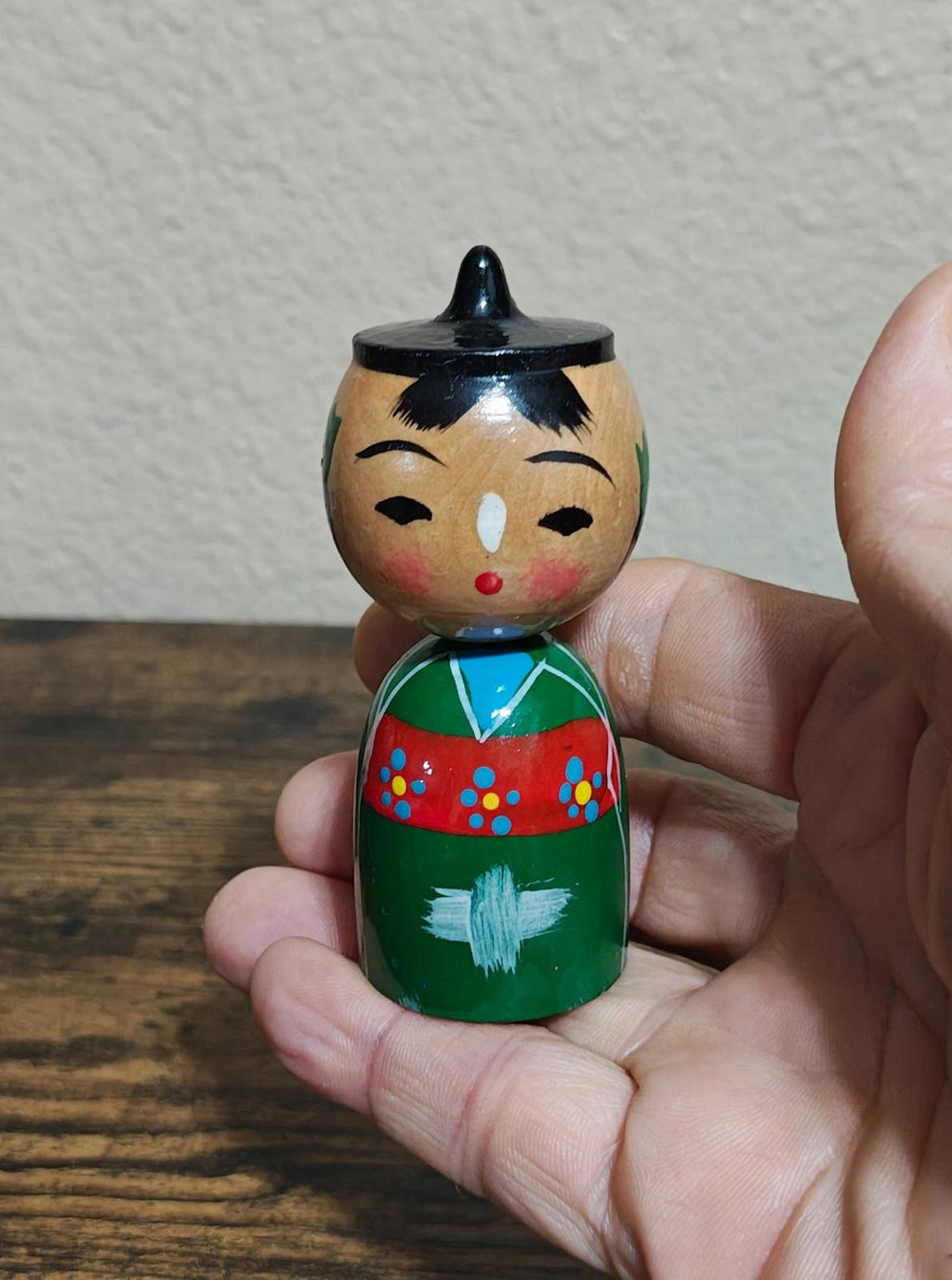Single Japanese kokeshi wooden doll in green dress held between fingers, red cheeks