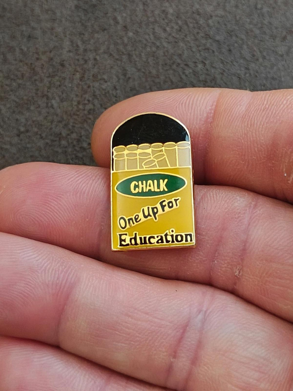 Close-up gold-tone enamel teacher pin with chalkboard text “CHALK / One Up For Education”
