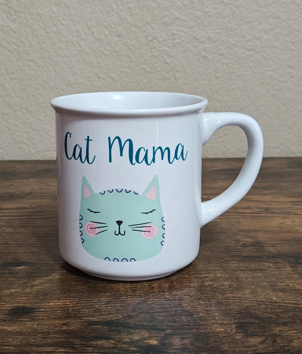 White ceramic mug with cat-face illustration and blue “Cat Mama” lettering