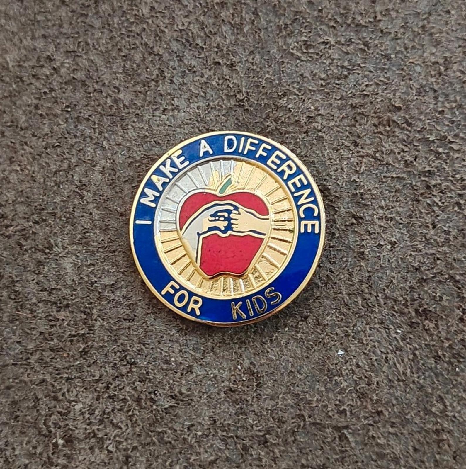 Gold-tone enamel pin showing blue “FOR KIDS” ring and apple/hand center design.