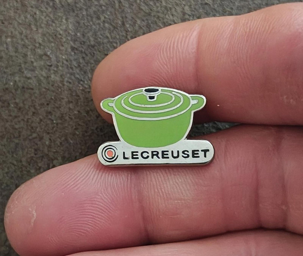 Le Creuset green mini cocotte enamel pin held on fingertip, front view