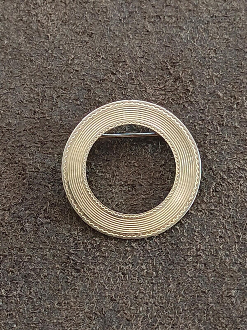Gold textured circular brooch, ring shape with central cutout on dark fabric background