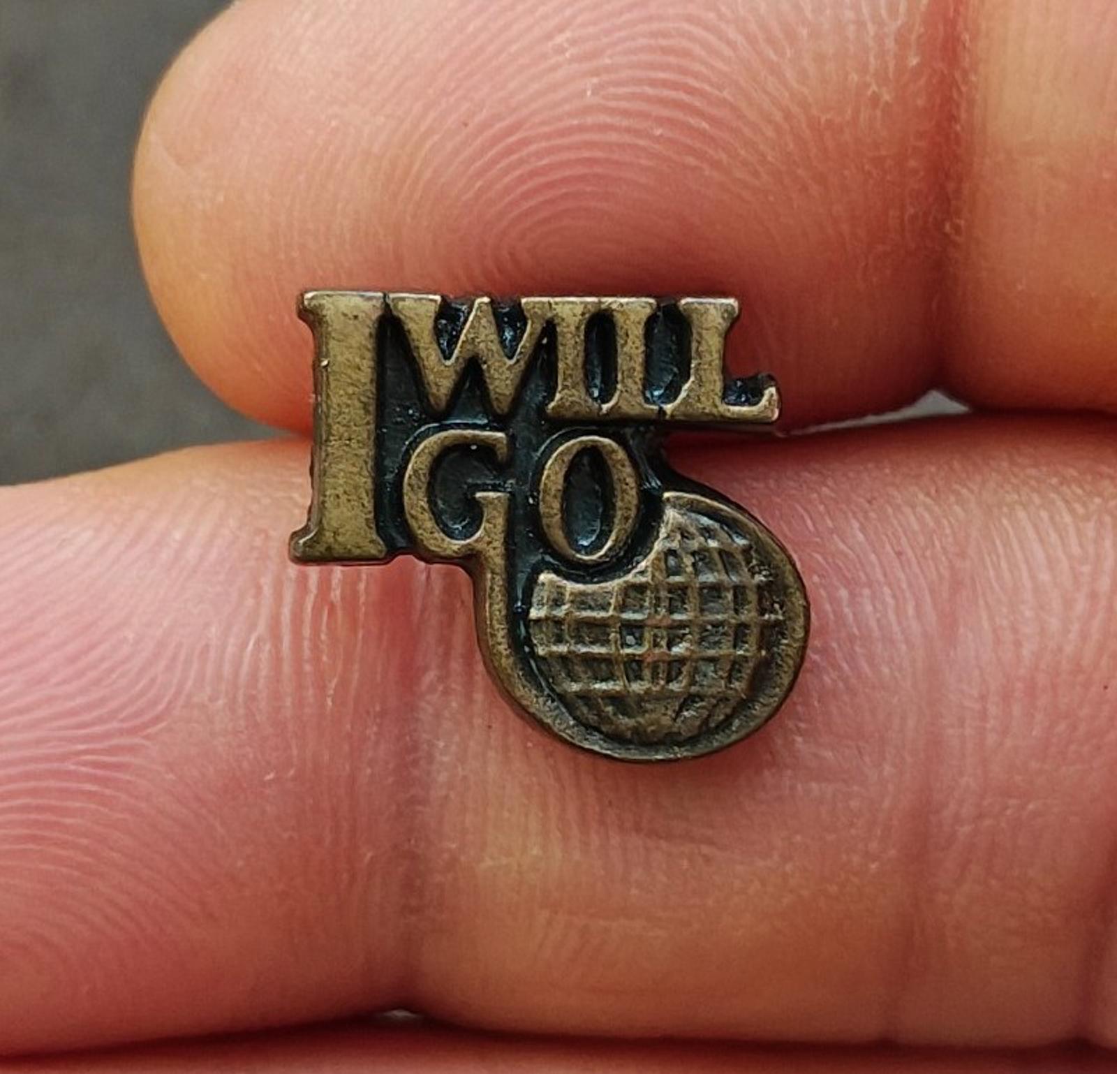 Gold tone "I WILL GO" lapel pin with globe-style emblem, close-up on fingertip
