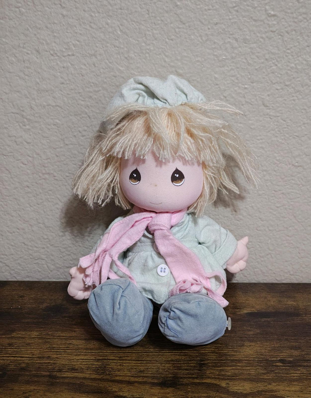 Beige plush doll with big eyes, mint-blue dress, pink scarf, sitting on table