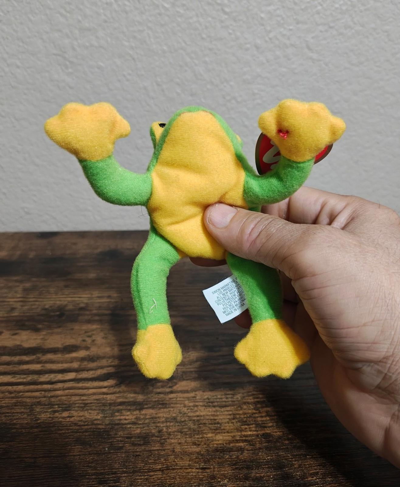 Green frog beanbag plush with yellow belly and raised arms, held in hand