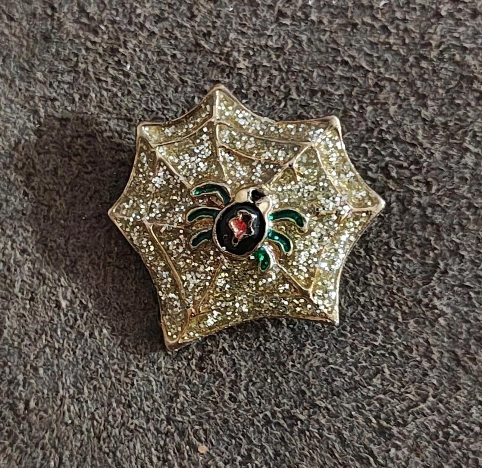 Gold spider web enamel pin on fabric, glitter texture, black spider body with red center, green legs
