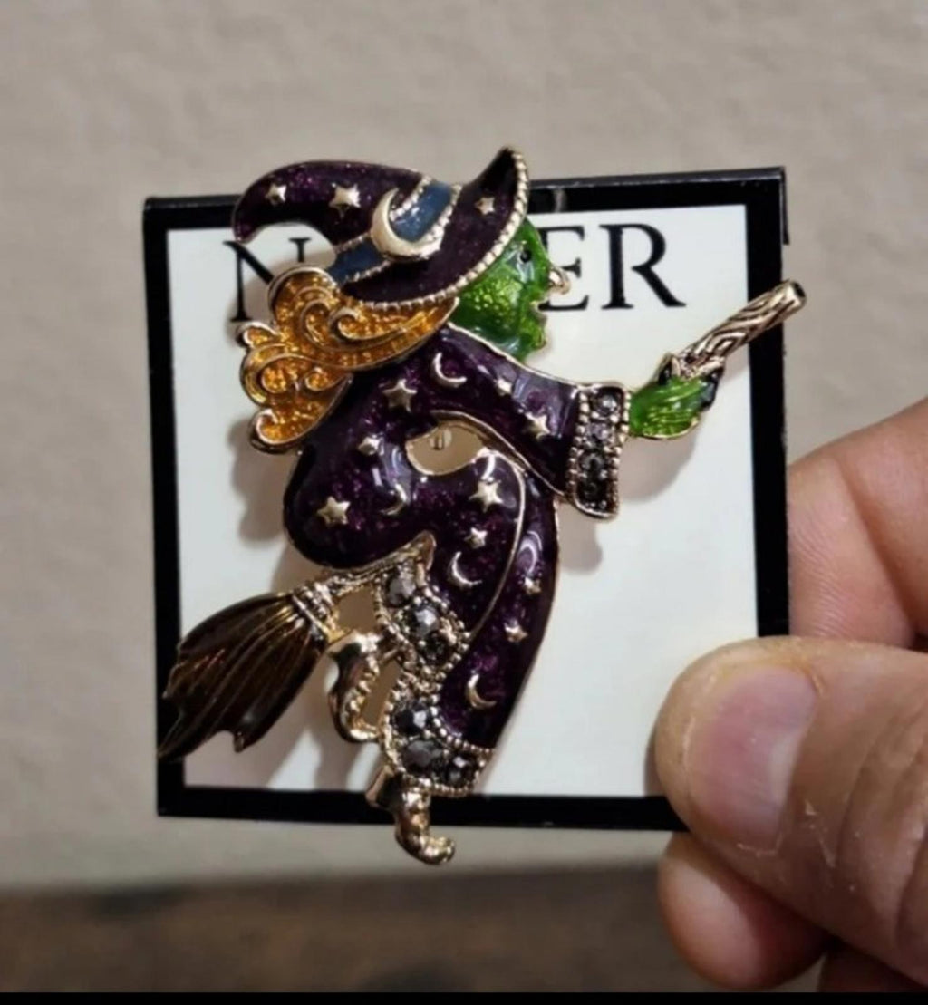 Gold-tone witch enamel pin brooch held by fingers, dark purple cloak and green broom