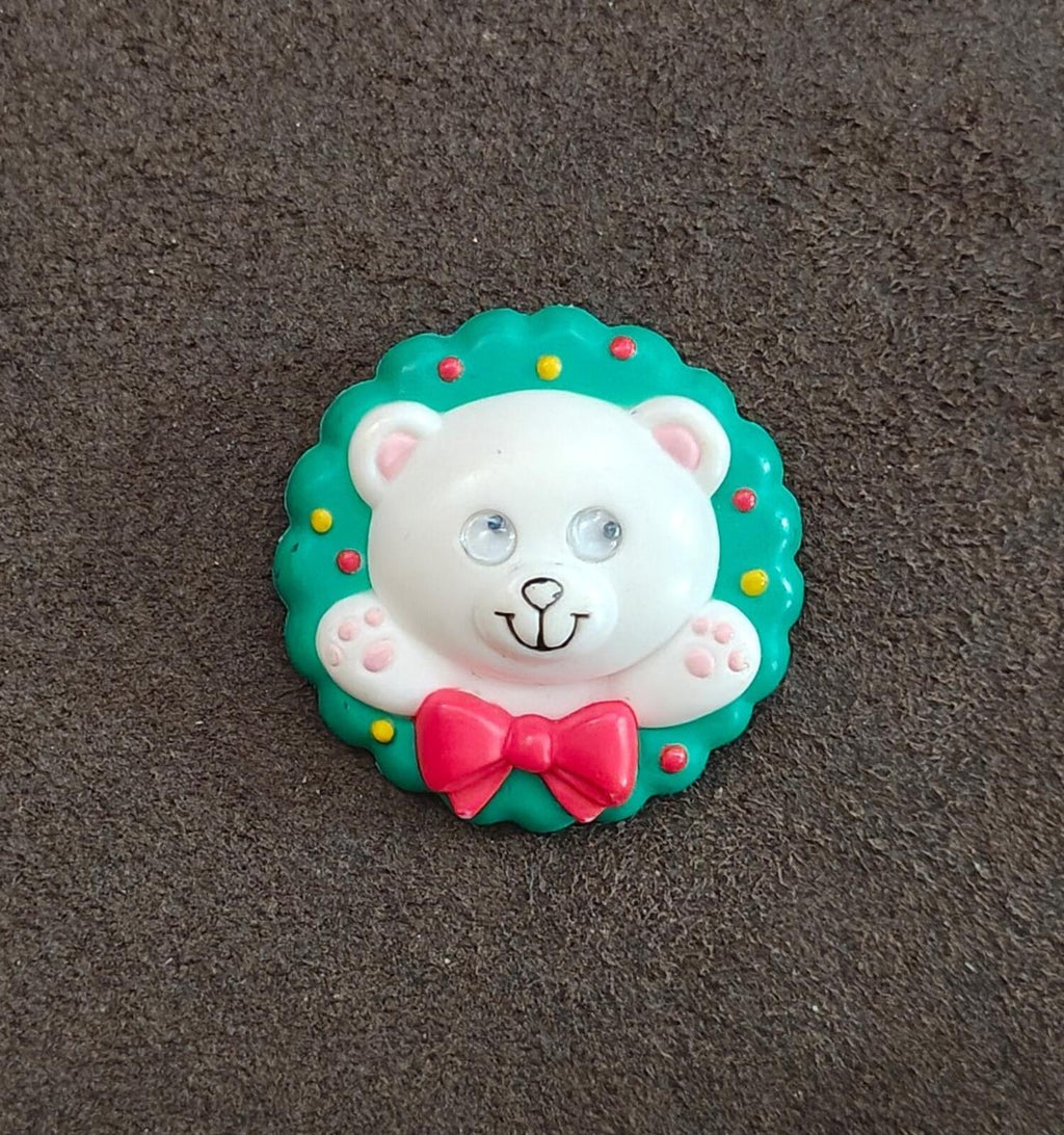 Green circular teddy bear wreath pin with white face and red bow on textured surface