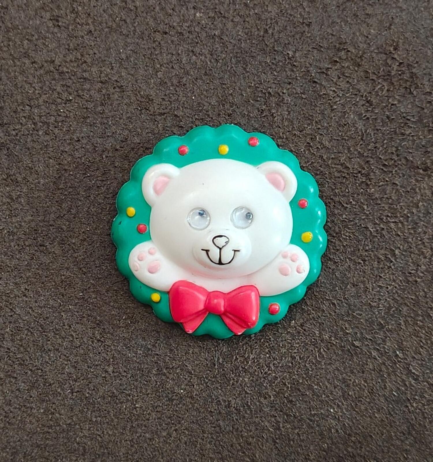 Green circular teddy bear wreath pin with white face and red bow on textured surface