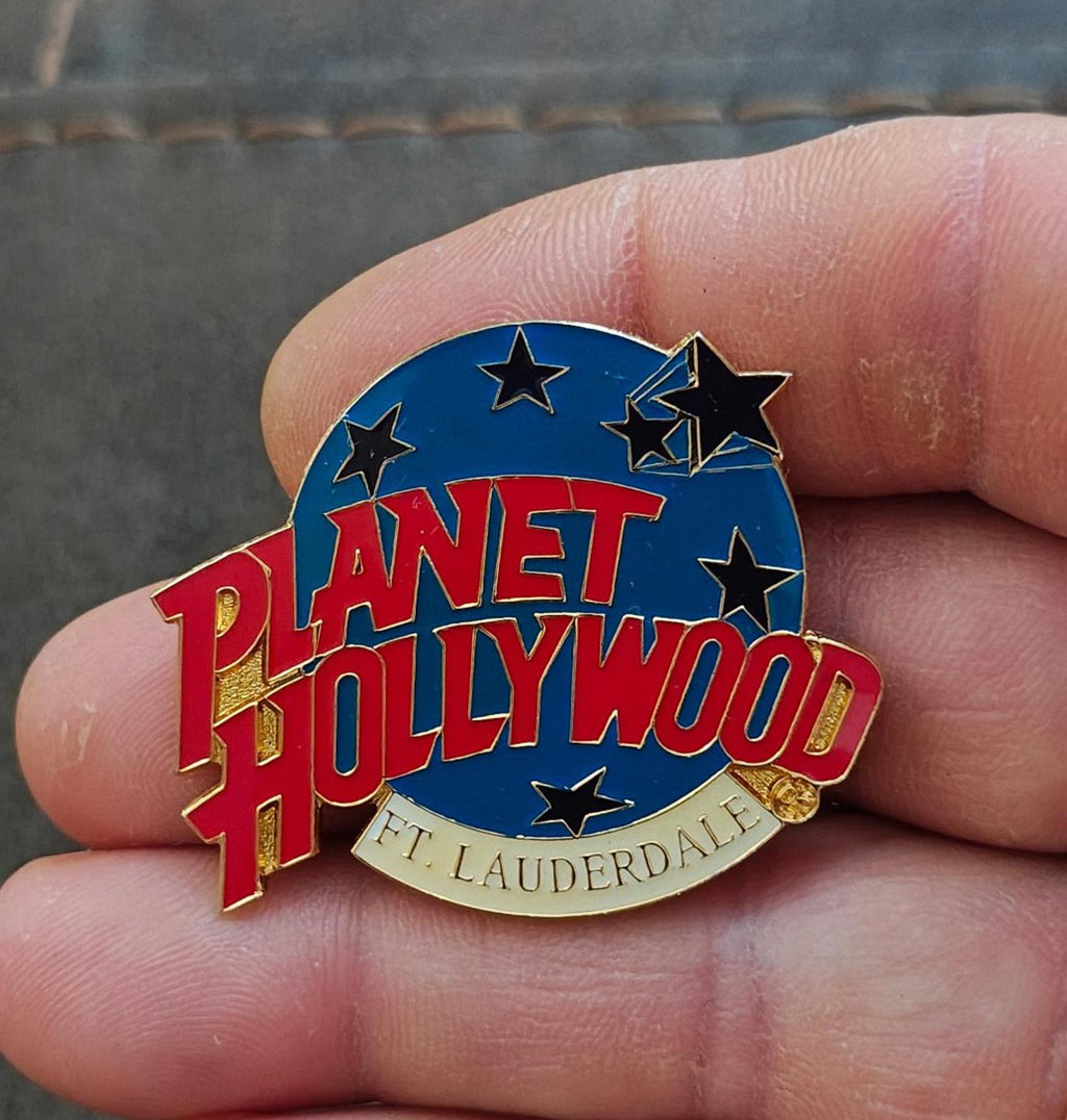 Blue enamel lapel pin with red Planet Hollywood lettering, star icons, gold edge.