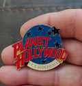 Blue enamel lapel pin with red Planet Hollywood lettering, star icons, gold edge.