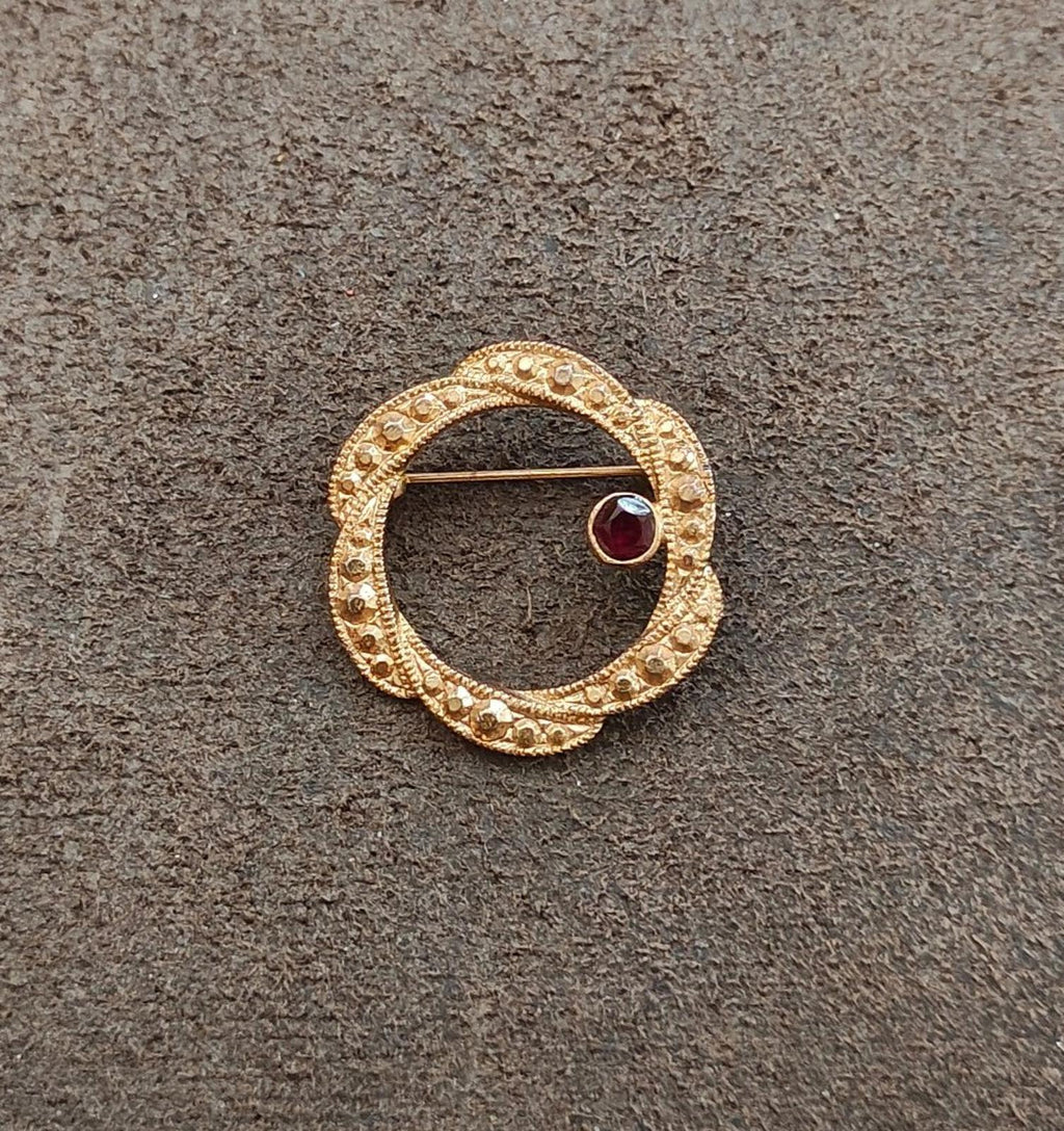 Gold tone circular brooch with dark red round garnet set in textured beaded frame.
