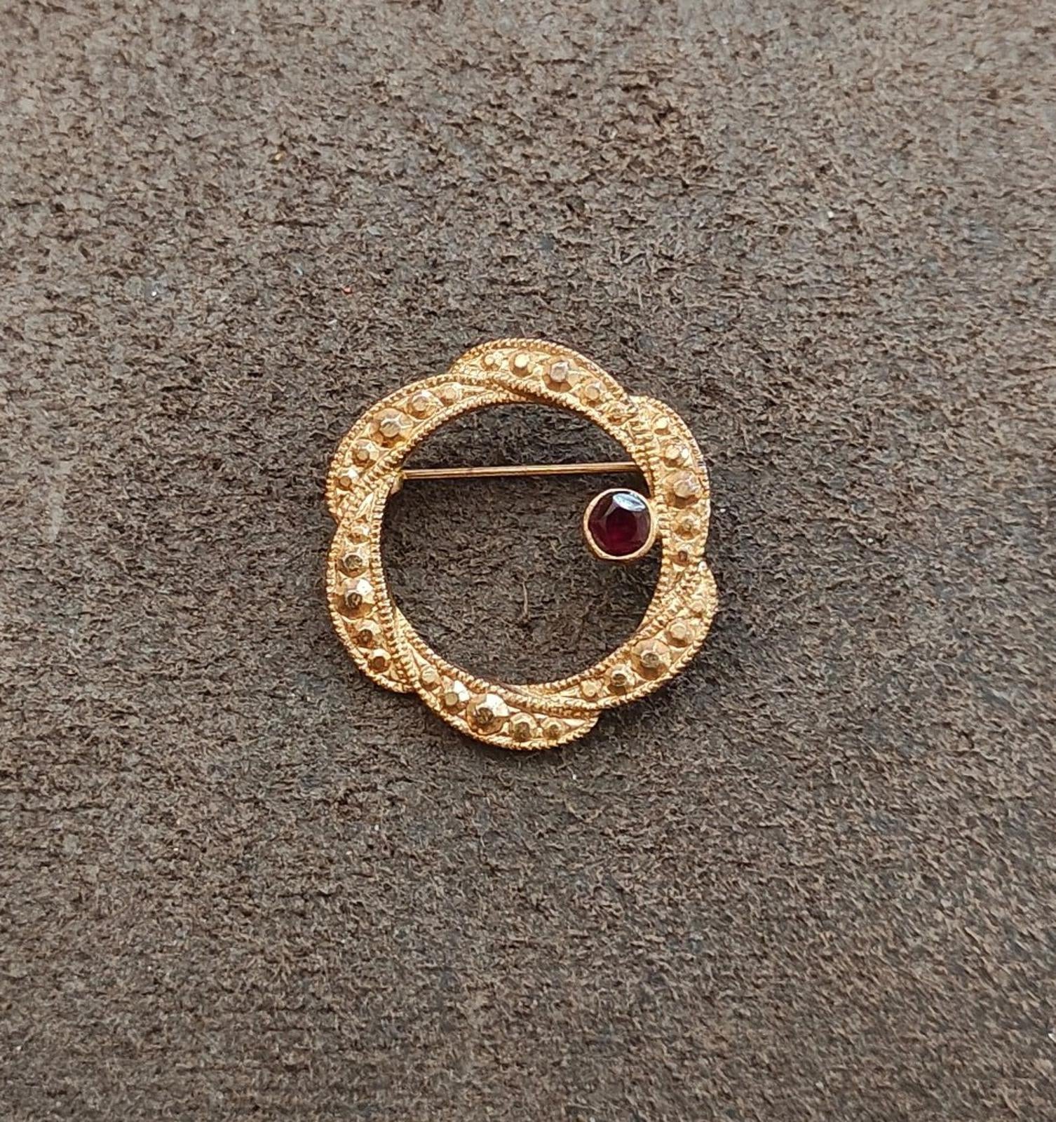 Gold tone circular brooch with dark red round garnet set in textured beaded frame.