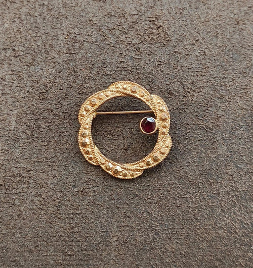 Gold tone circular brooch with dark red round garnet set in textured beaded frame.