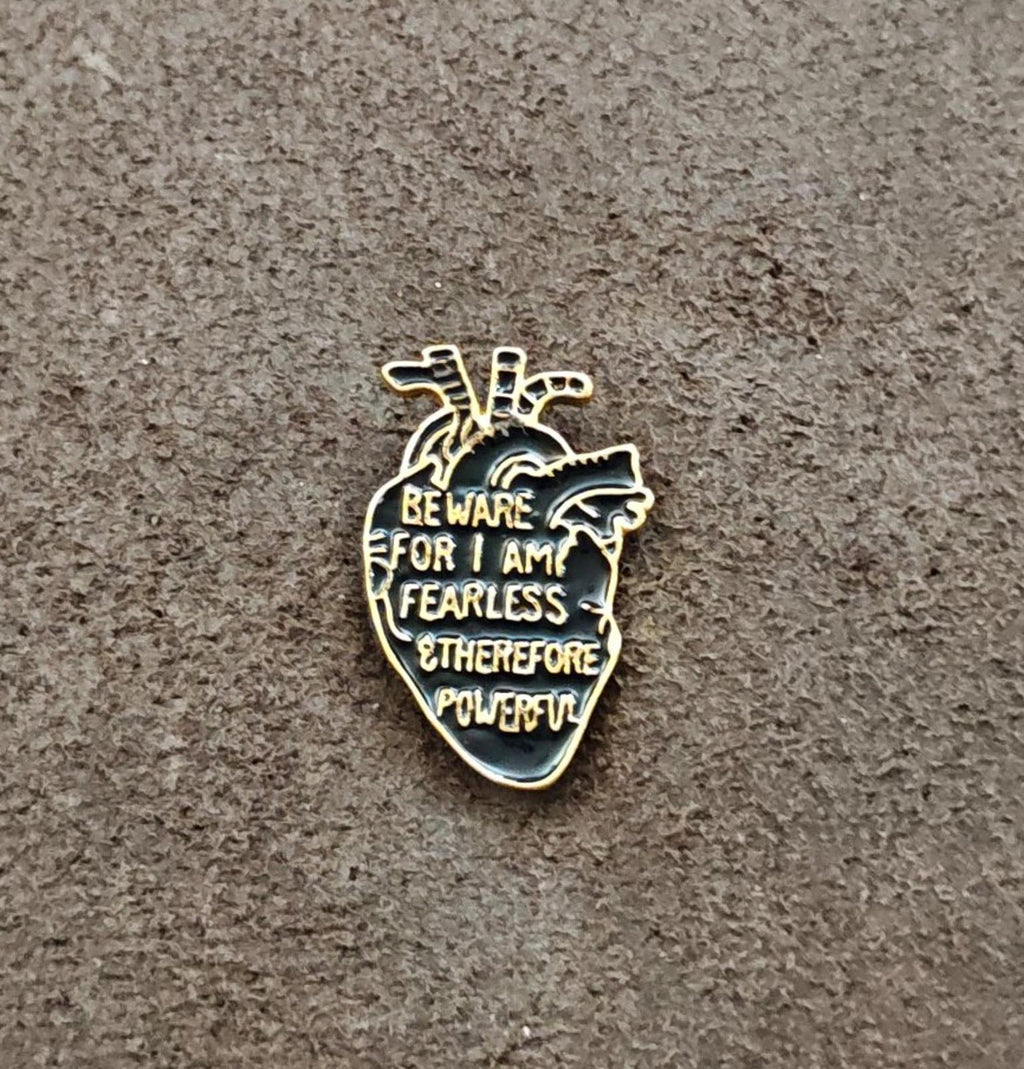 Gold-tone anatomical heart enamel pin with black enamel and warning text on gray surface