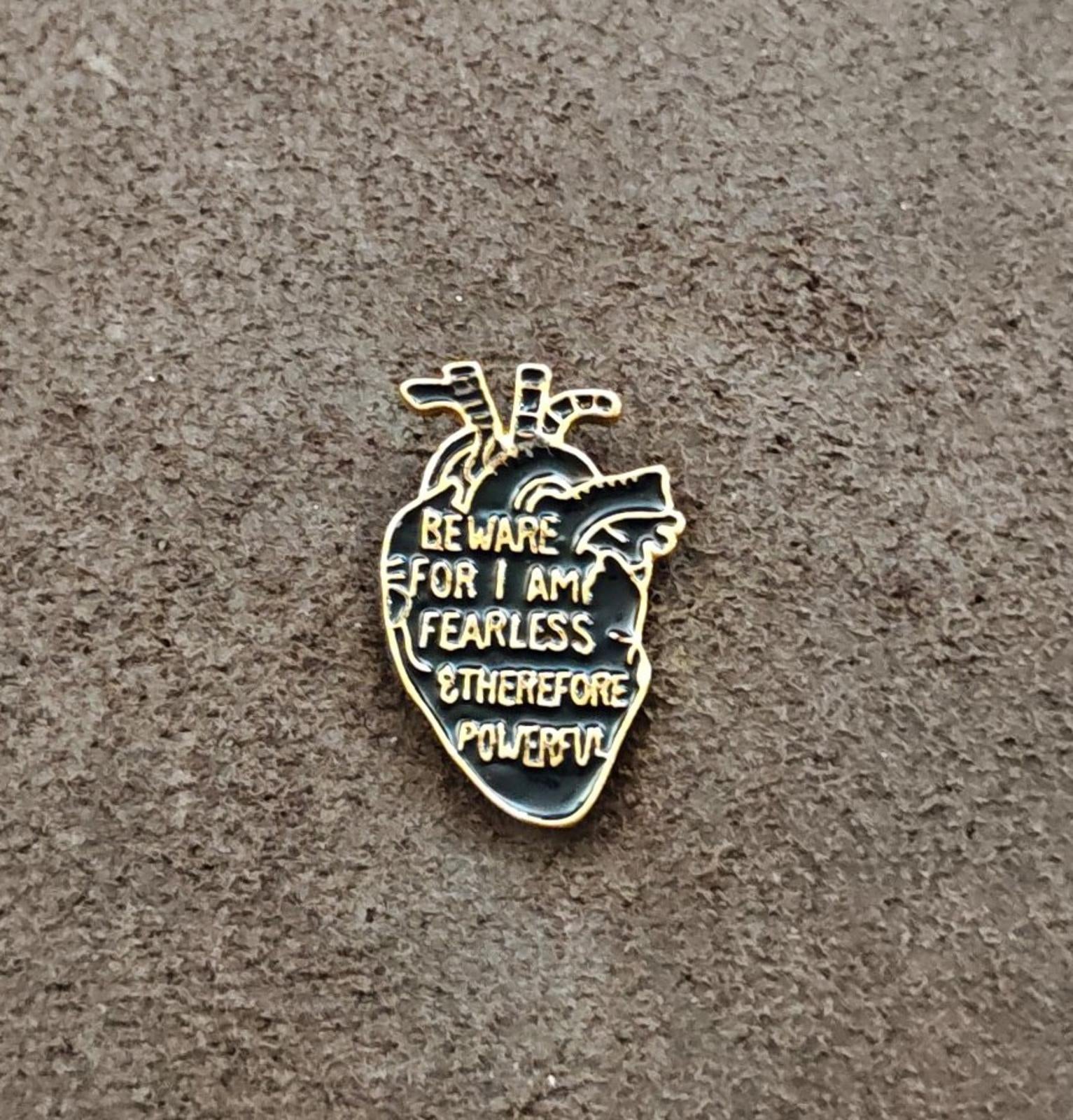 Gold-tone anatomical heart enamel pin with black enamel and warning text on gray surface
