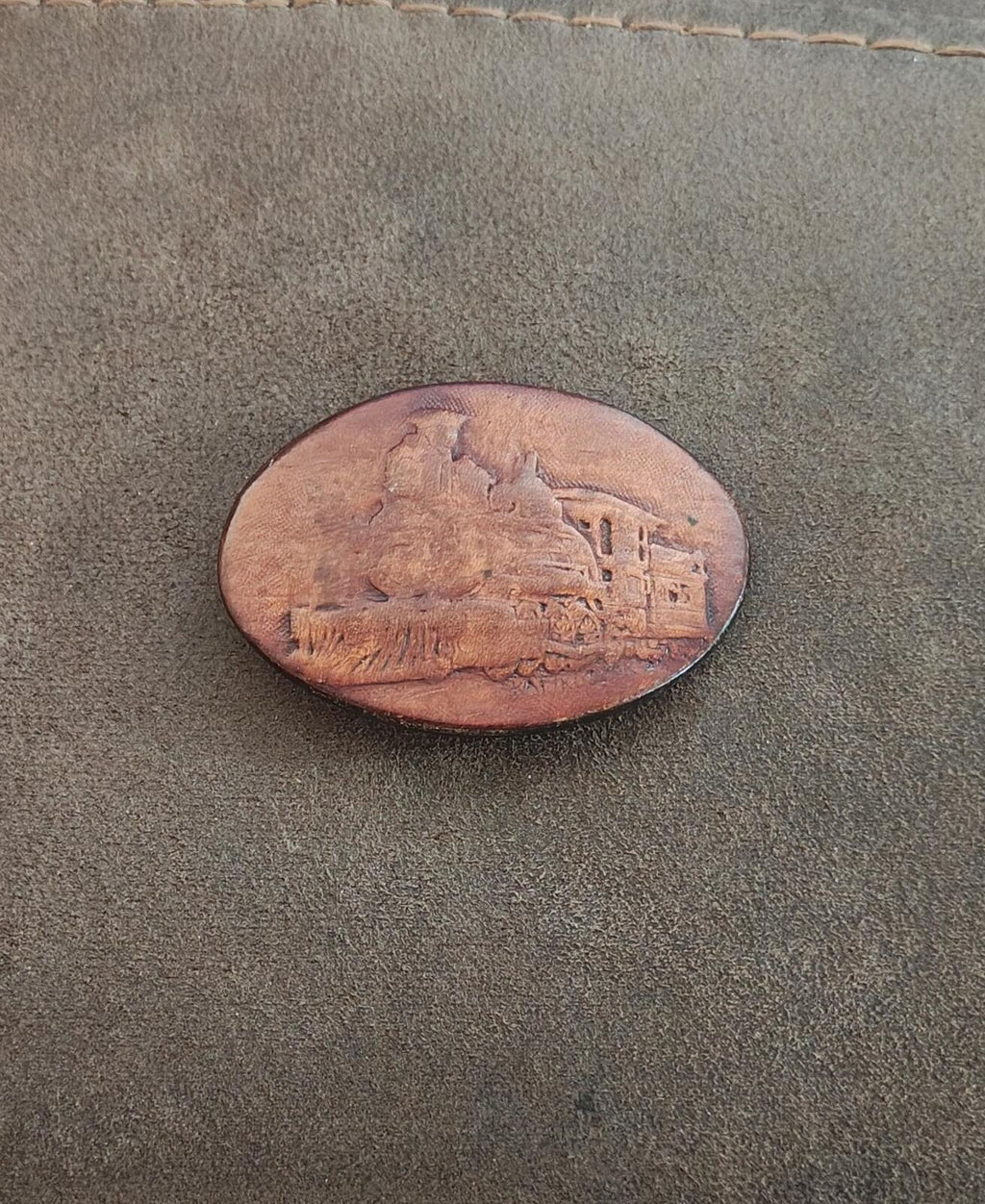 Brown oval leather belt buckle lying flat with embossed steam locomotive