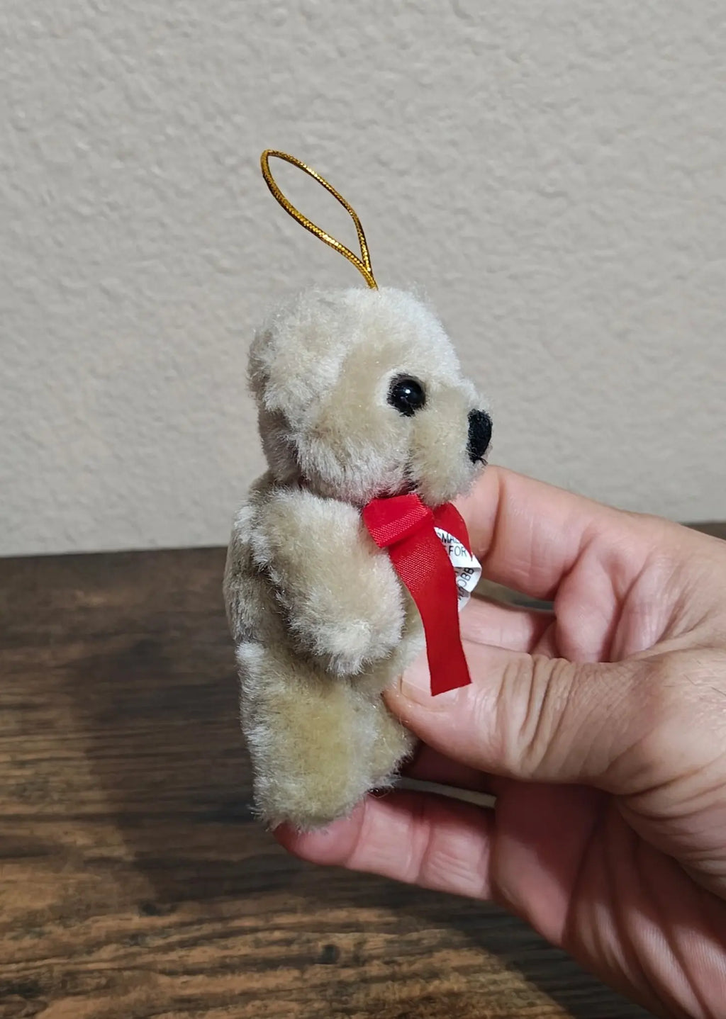 Hand holding beige plush teddy bear ornament with red bow and gold hanging loop cord