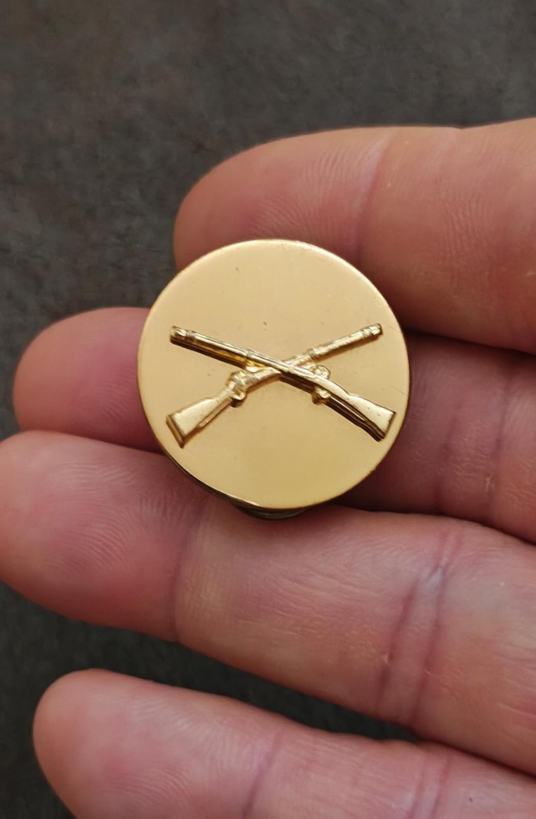 Gold circular brass pin with crossed rifles emblem, close-up front