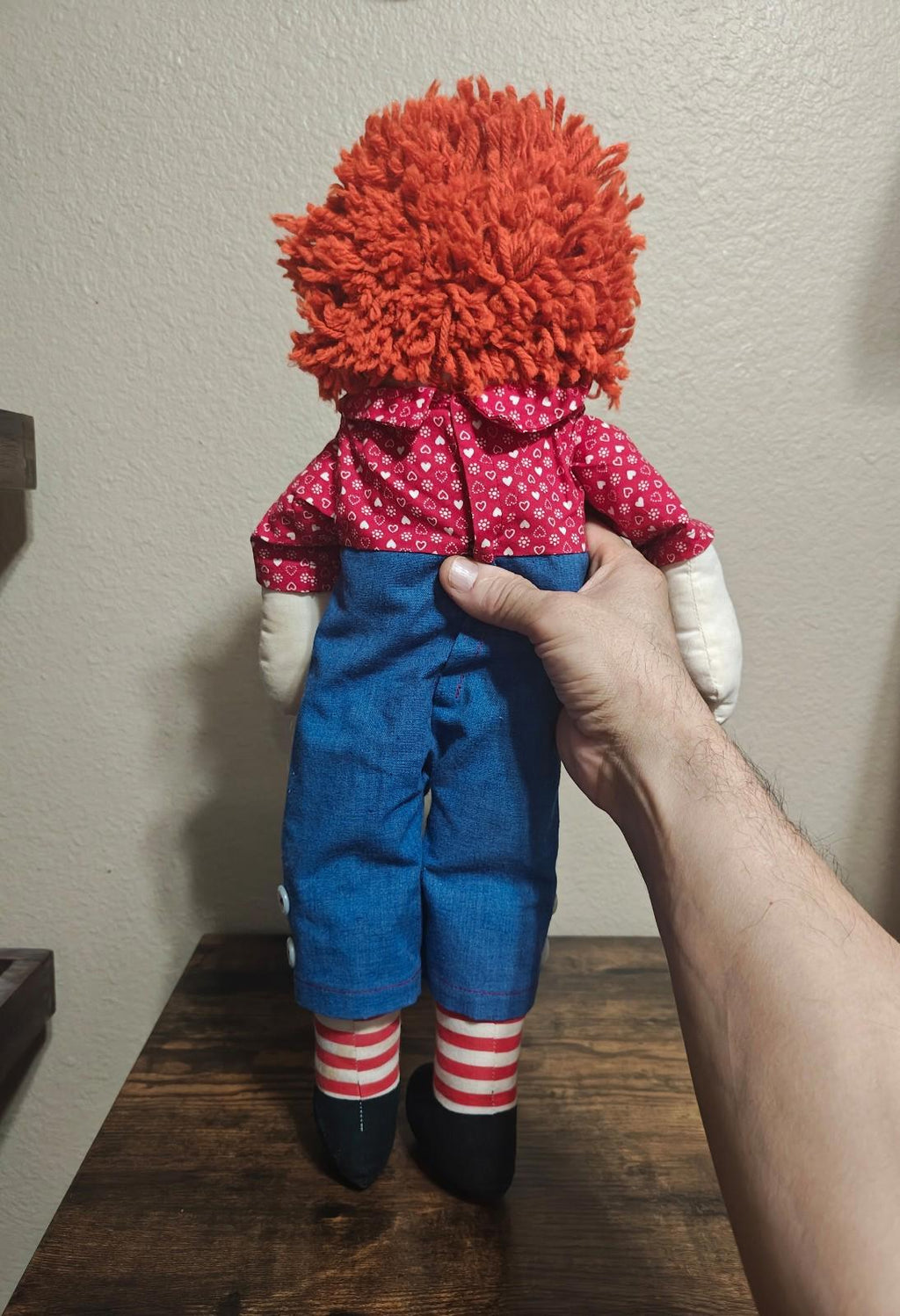 Handmade rag doll back view with orange yarn hair and red heart-pattern shirt over blue overalls.
