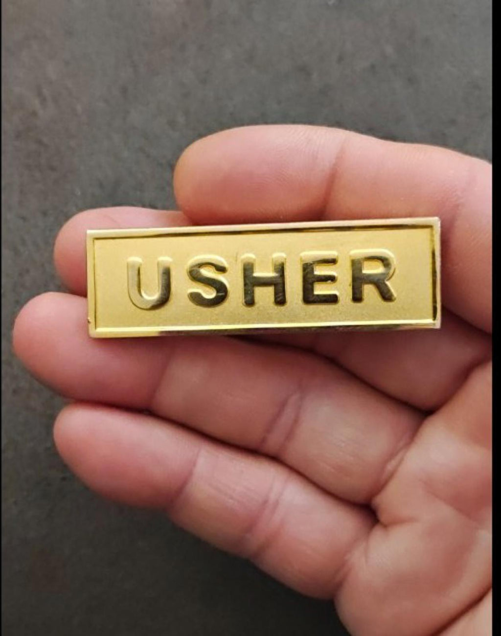 Gold tone usher badge pin held in hand, embossed USHER lettering