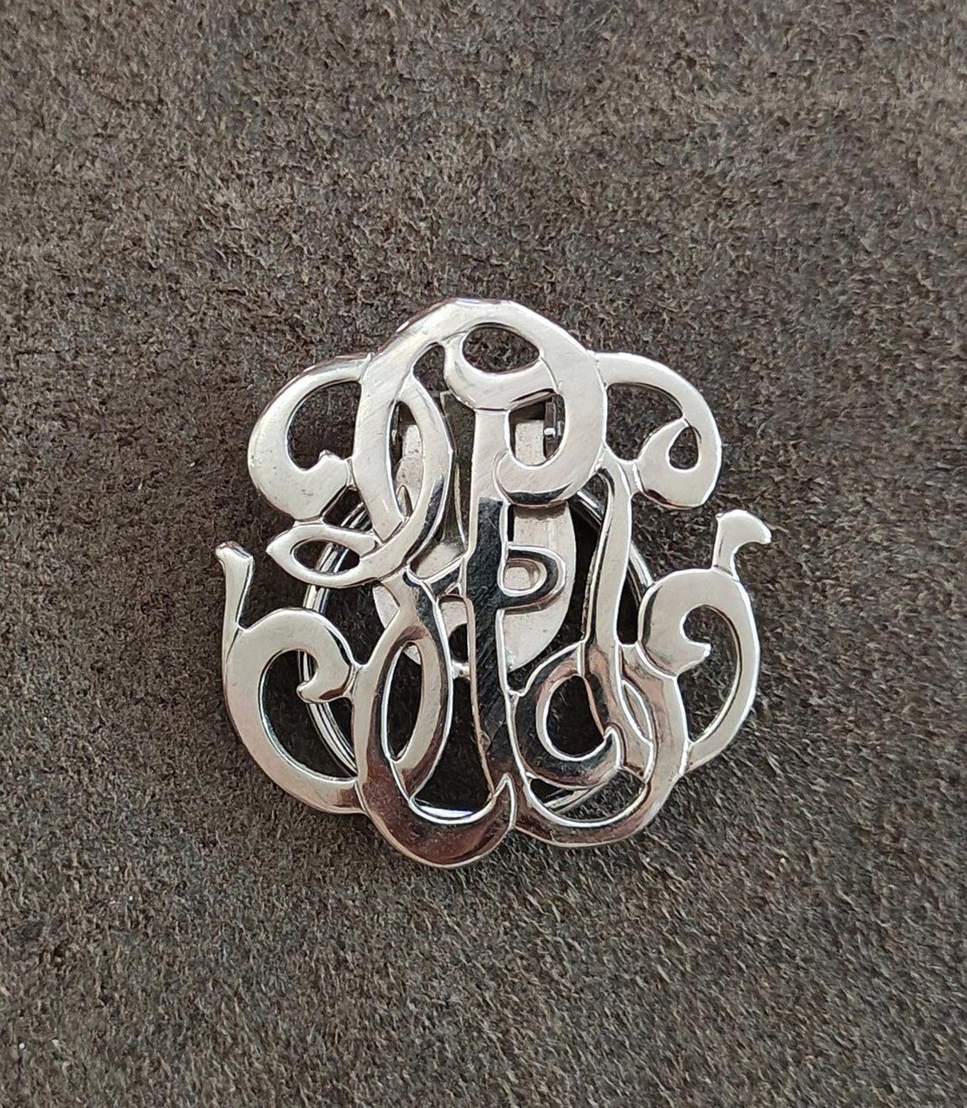 Silver-tone monogram scarf clip, close-up of polished ornate monogram