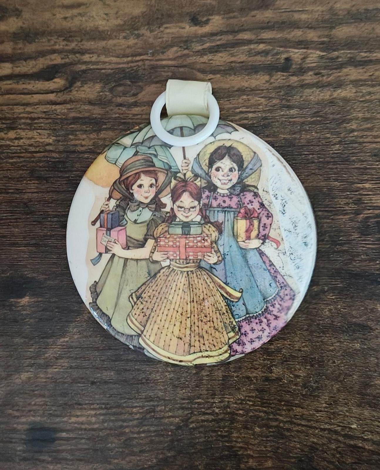 Round hanging ornament with colorful illustrated girls and a small toy cart, cream top loop